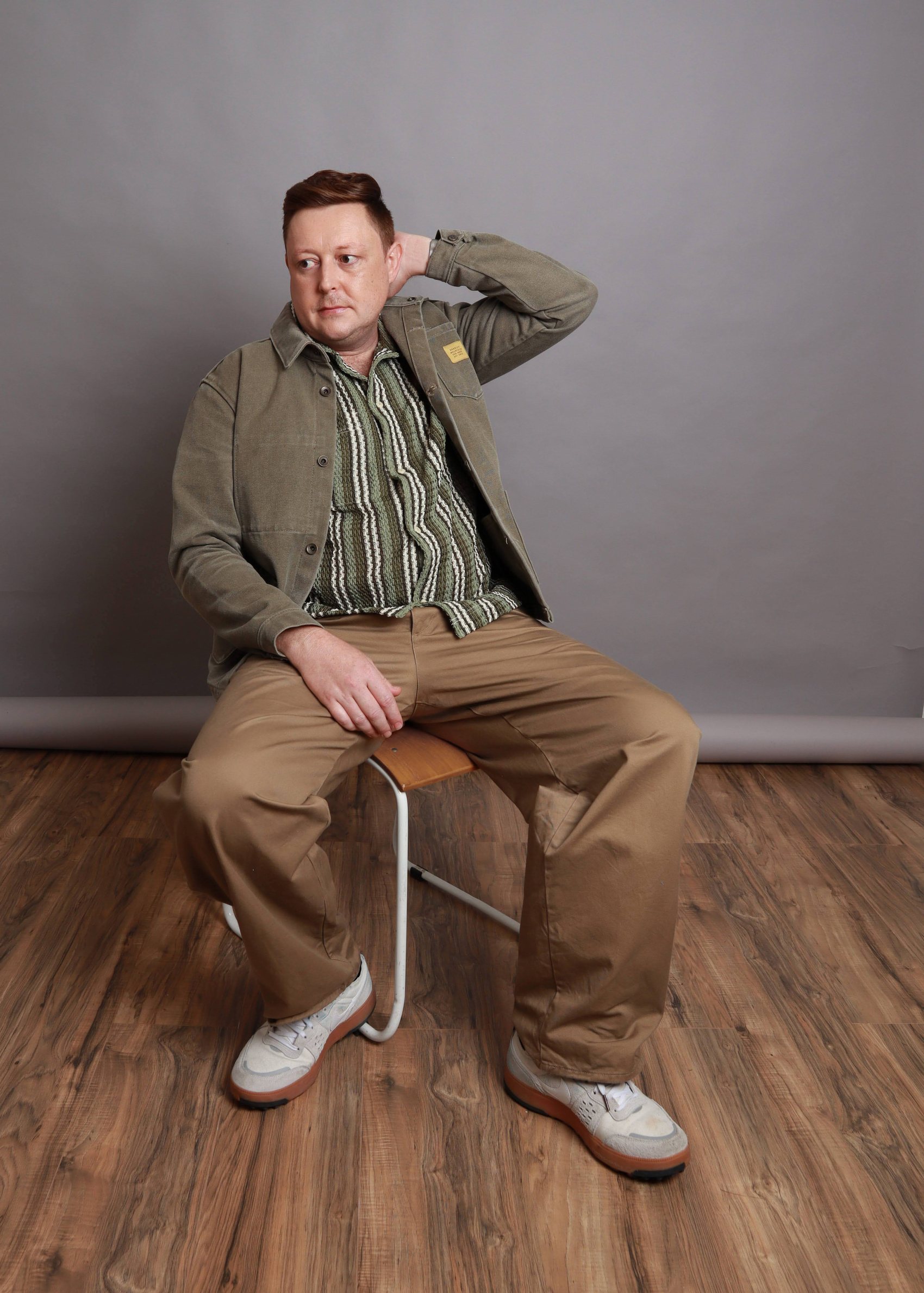 Cameron seated on a stool in an olive overshirt and striped knit, textured side-part haircut, Bang Bang Stone grooming work