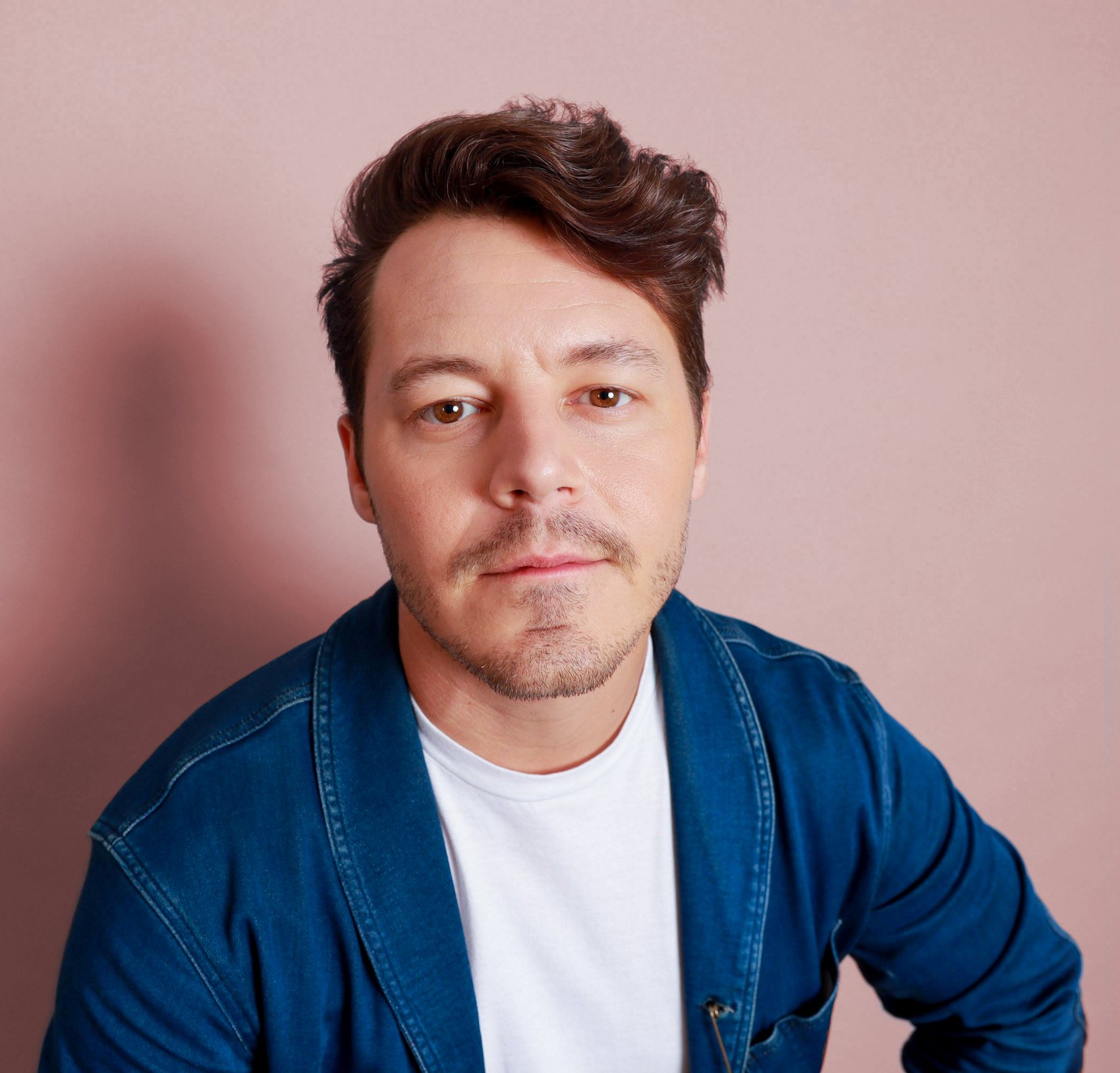 Portrait against a pink backdrop in a denim shirt over a white tee, tousled side-part hair, Bang Bang Stone men's grooming