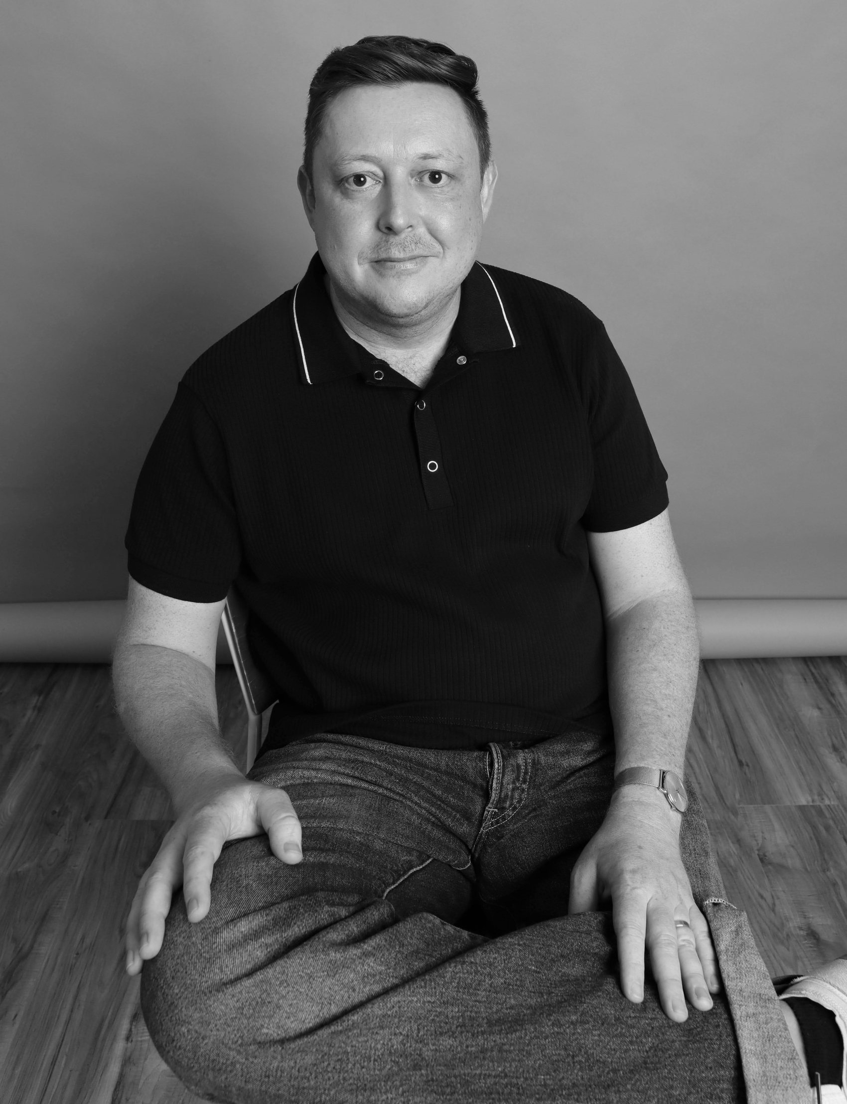 Black-and-white seated portrait in a black polo, cropped textured haircut, Bang Bang Stone men's grooming