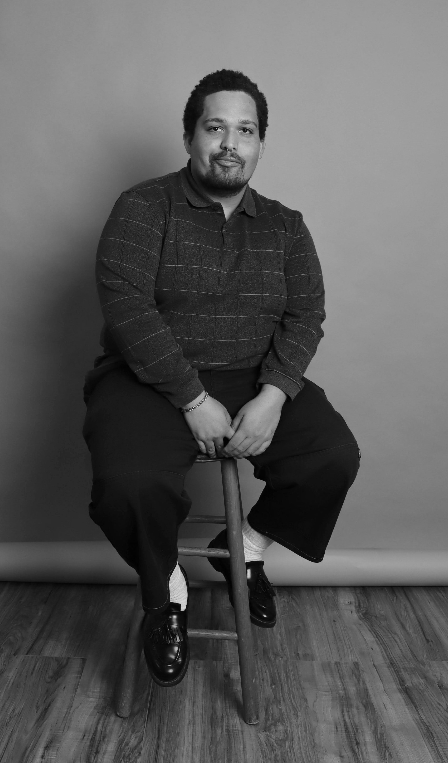 Black-and-white seated portrait in a striped polo with arms resting on the knees, Bang Bang Stone men's grooming