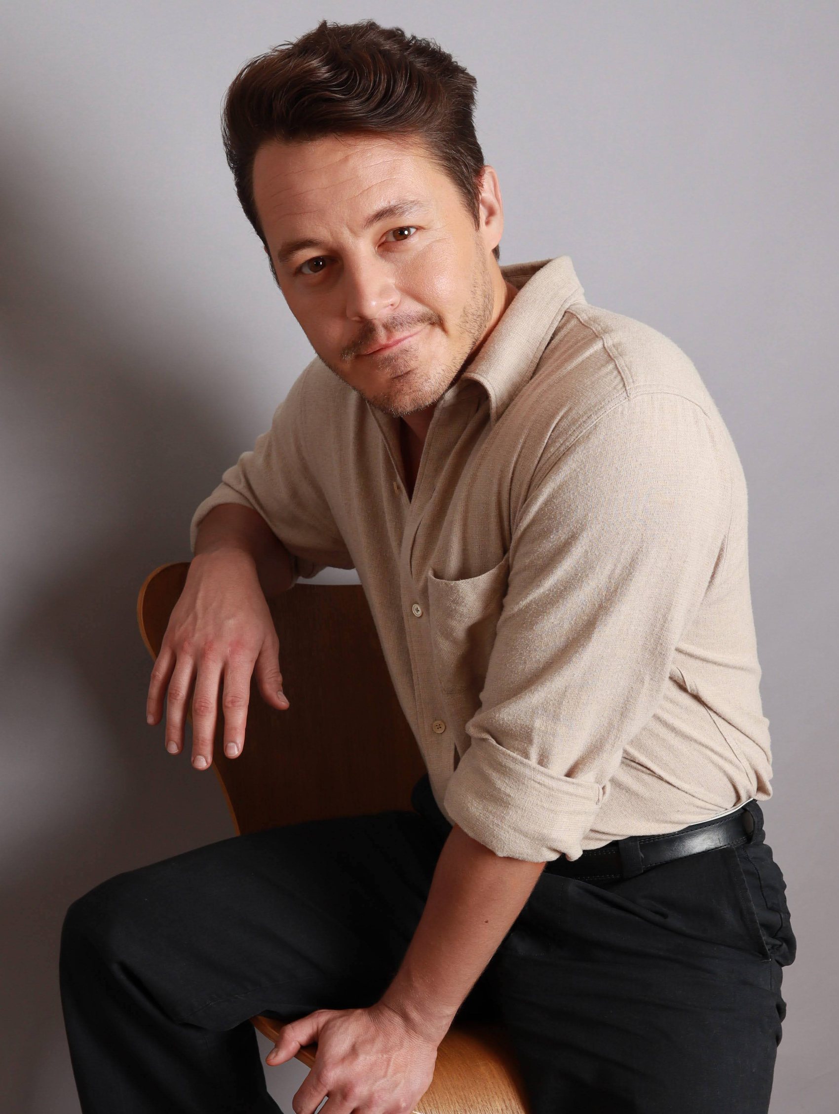 Seated portrait in a beige button-up with a relaxed side part, leaning on a wooden chair back, Bang Bang Stone grooming work