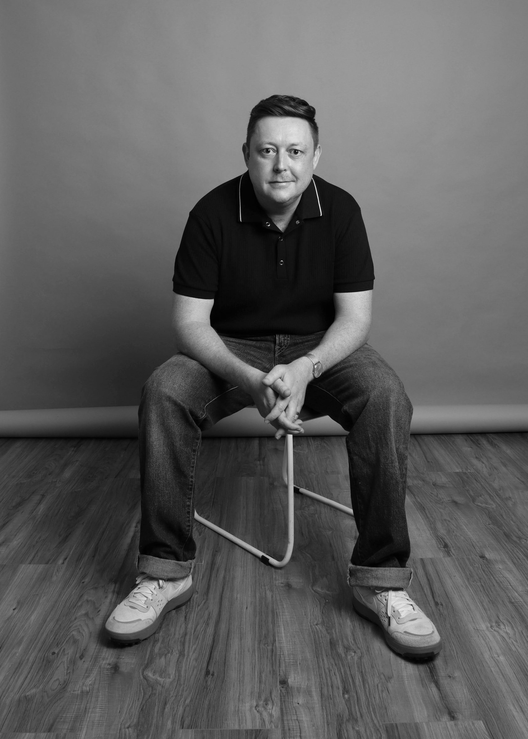 Black-and-white seated portrait in a black polo with hands clasped, cropped haircut, Bang Bang Stone men's grooming
