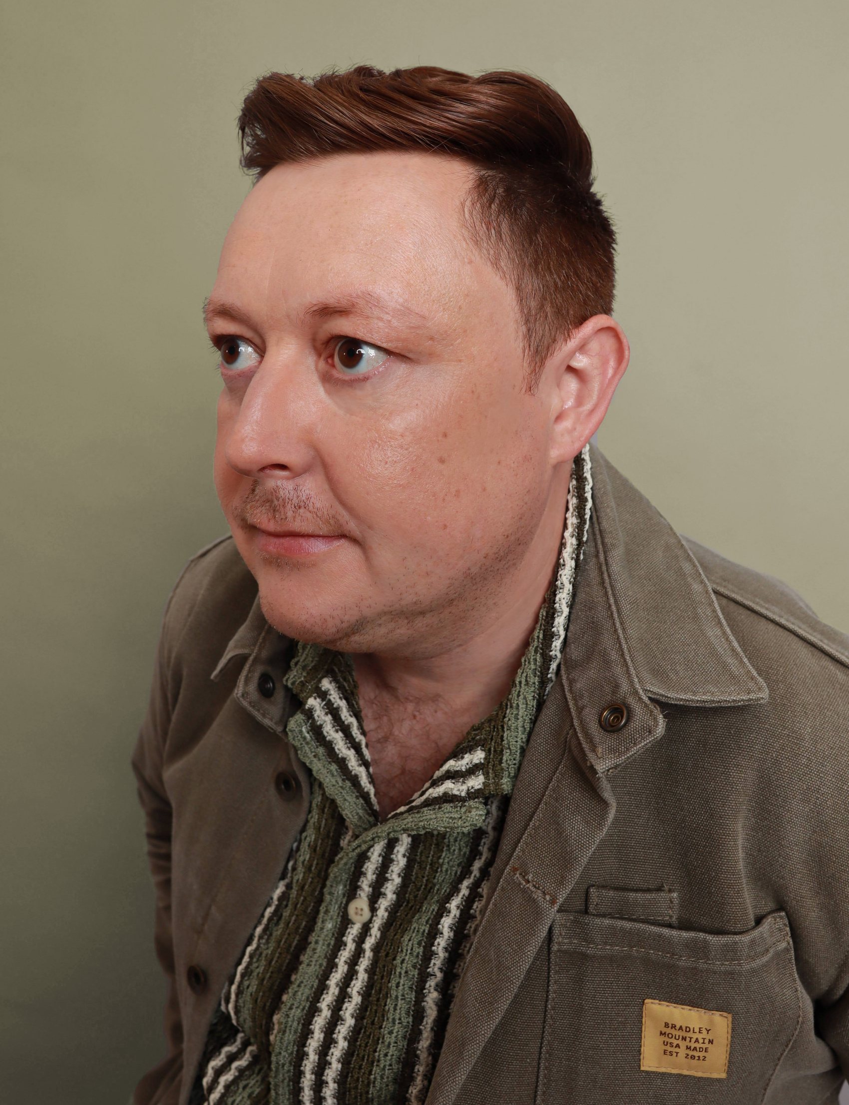 Cameron in profile wearing a striped scarf and olive jacket against a sage-green backdrop, auburn hair, Bang Bang Stone grooming work