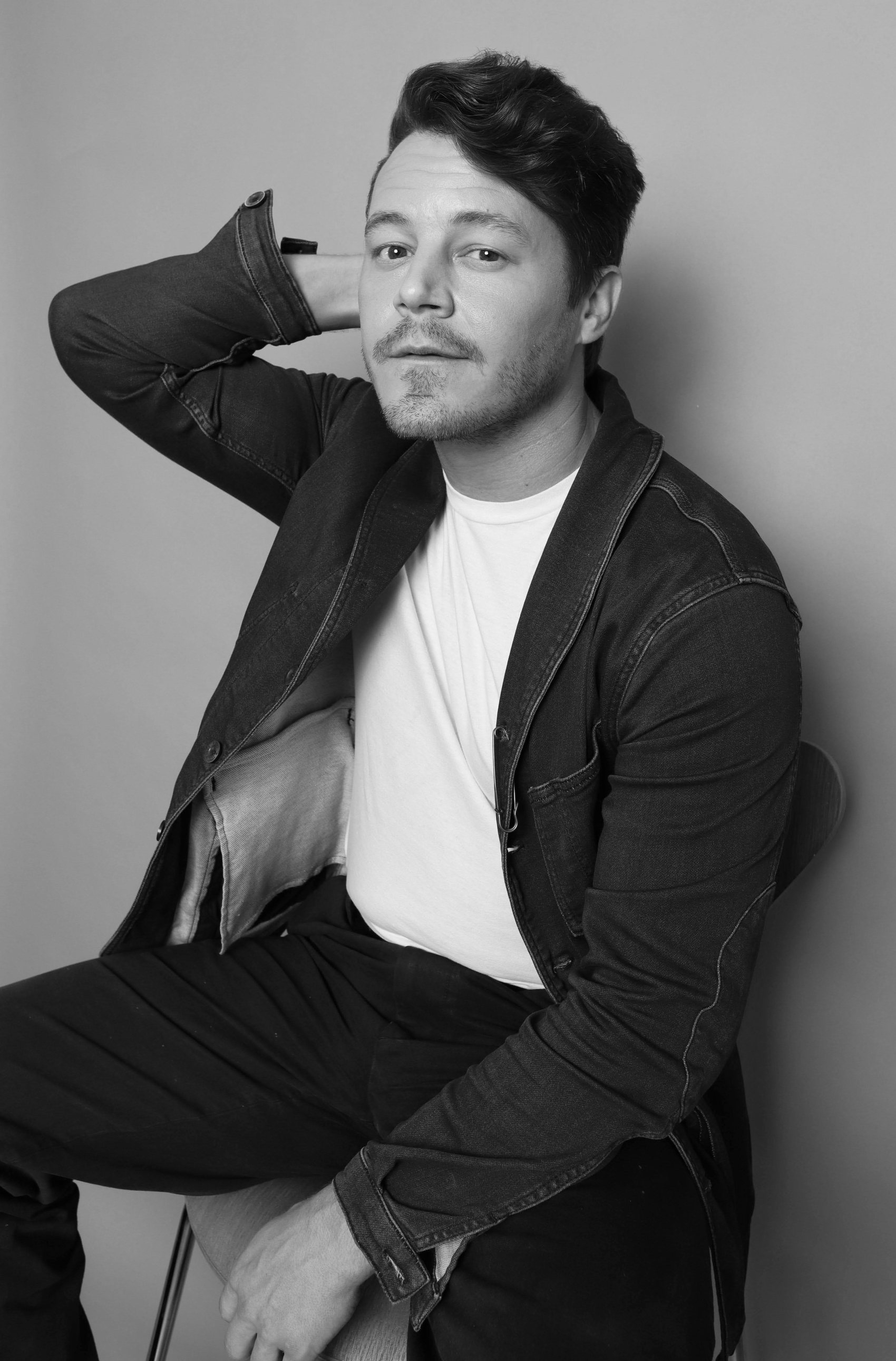 Black-and-white seated side portrait in a denim jacket over a white tee, hand resting behind the head, Bang Bang Stone men's grooming