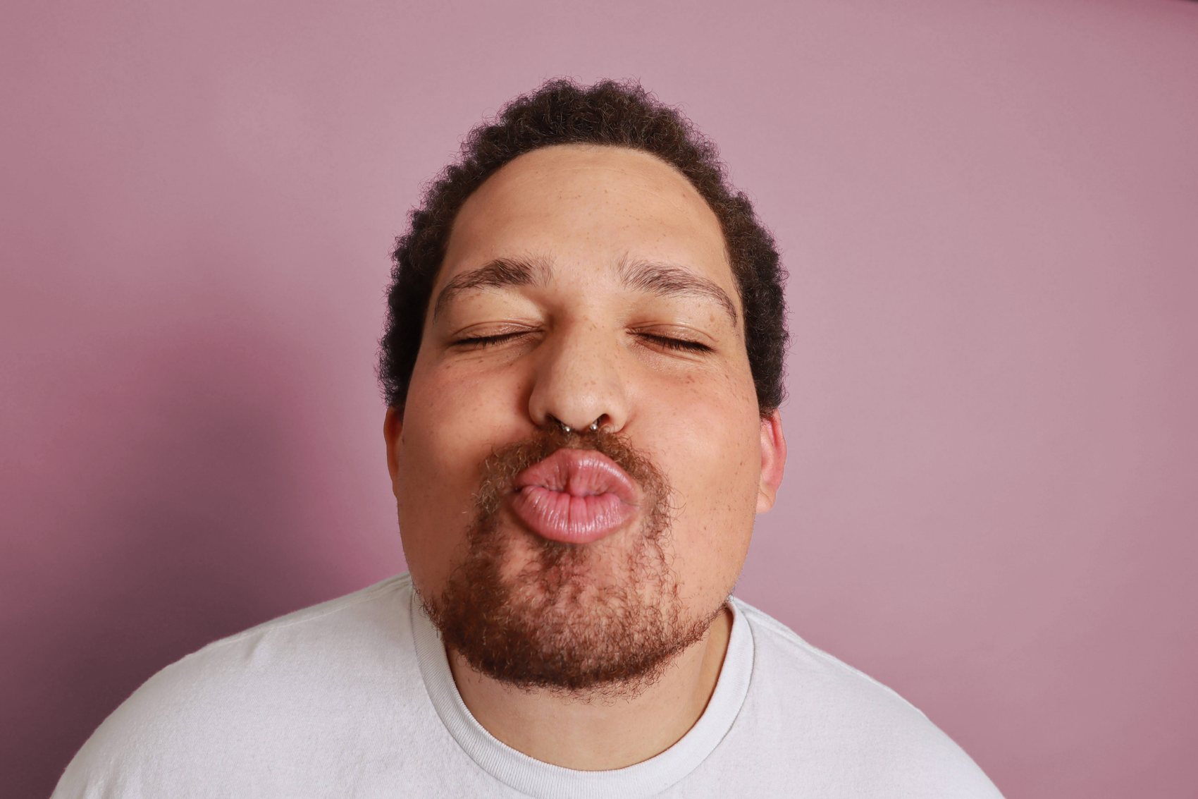 Playful close-up against a pink backdrop, eyes closed with a pursed expression, groomed beard, Bang Bang Stone men's grooming